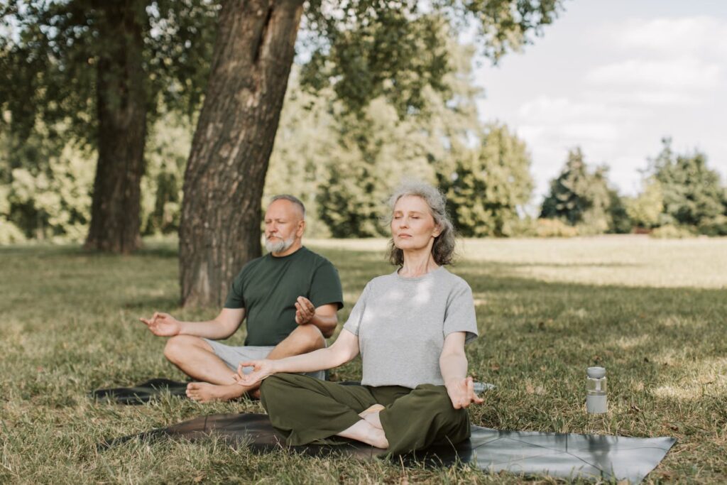 Elderly couple sitting cross-legged on yoga mats in park, meditating under shade of large trees.