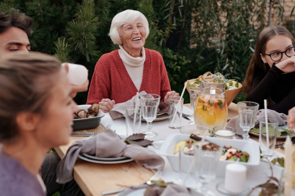 An elderly woman laughing and enjoying a meal outdoors with family and friends, gathered around a table filled with food and drinks.