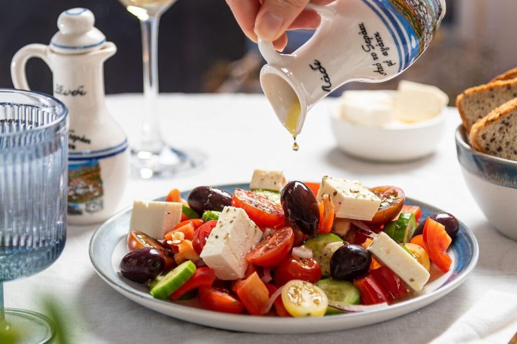 Hand drizzling olive oil onto vibrant Greek salad, topped with feta cheese, olives, tomatoes, cucumbers, and bell peppers.