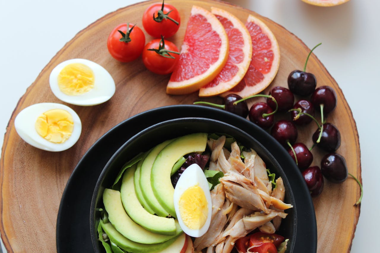 Healthy meal high in ellagic acid, consisting of avocado slices, shredded chicken, and boiled egg in a black bowl, surrounded by fresh fruits.