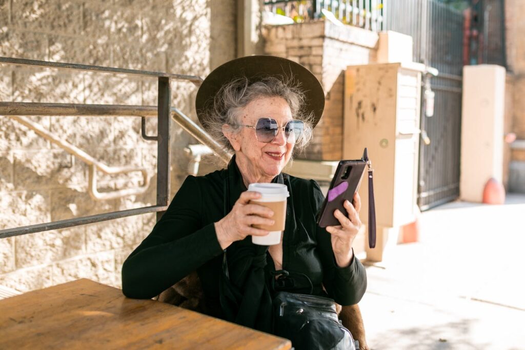 Older woman sitting at outdoor café, smiling as she looks at smartphone while holding a cup of coffee.