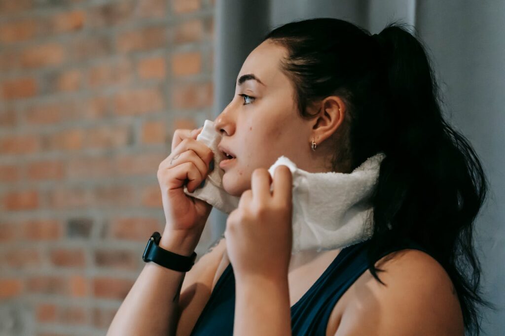Woman with dark hair in ponytail wipes face with a towel after workout, appearing slightly out of breath.