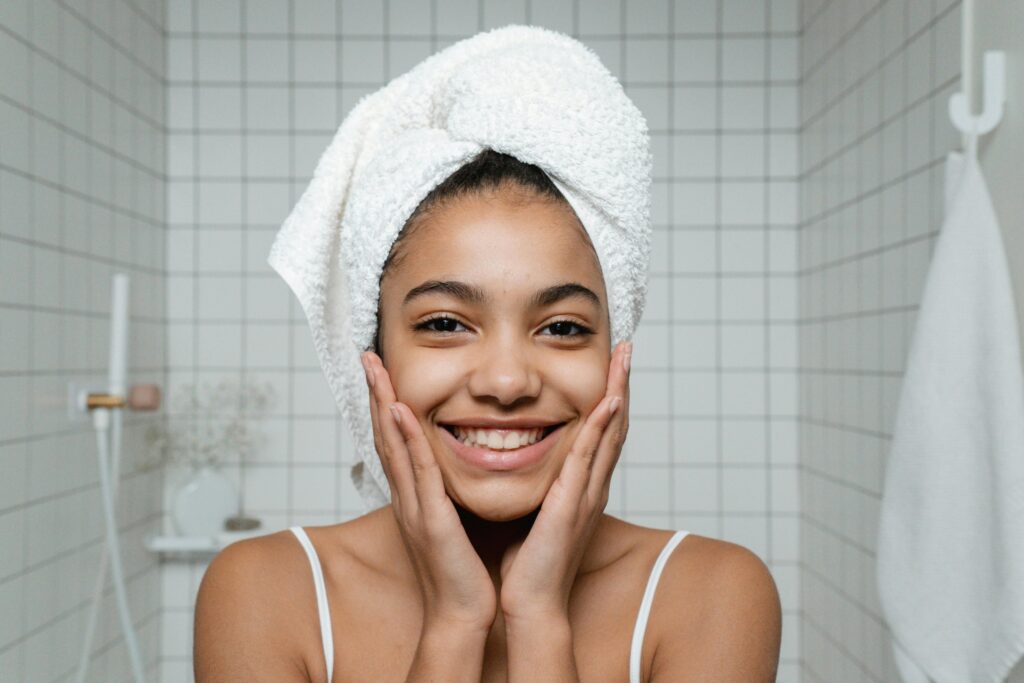 Young woman smiling confidently while standing in a tiled bathroom, with a towel wrapped around hair, showcasing clear and healthy skin.
