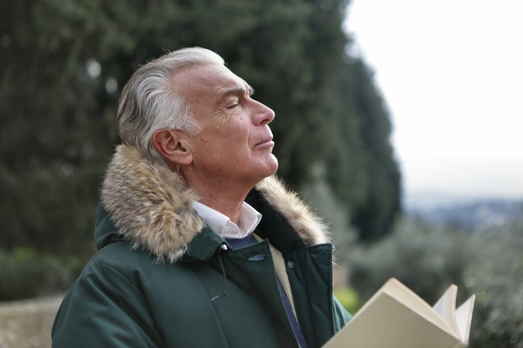 A man standing outside with eyes closed, enjoying the fresh air, while holding an open book.