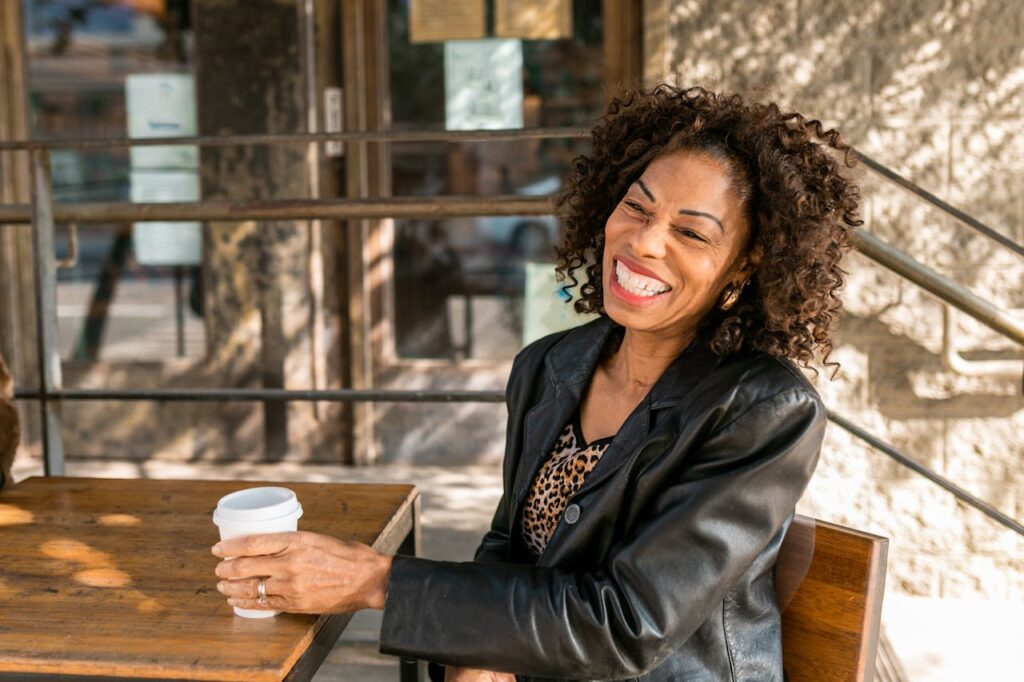 A woman with curly hair and a leather jacket sits at a table, smiling while holding a cup of coffee.