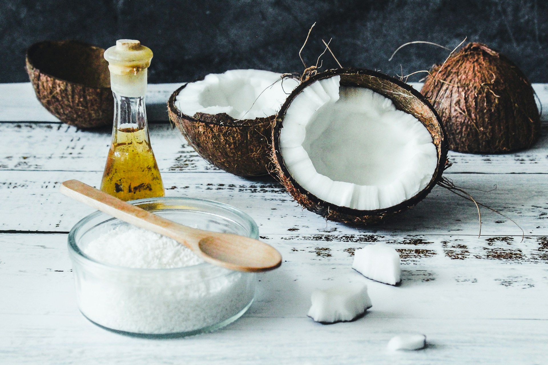Half of a cracked coconut, coconut oil in a glass bottle, and a bowl of shredded coconut with a wooden spoon.