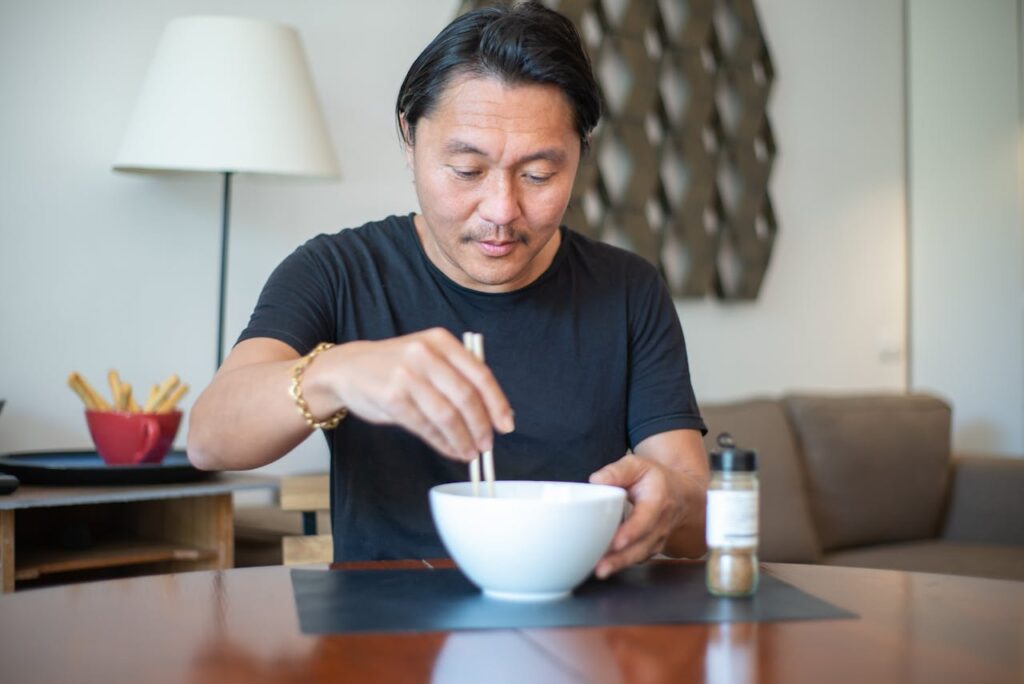 Man using chopsticks to eat from bowl while sitting at table in a cozy indoor setting.
