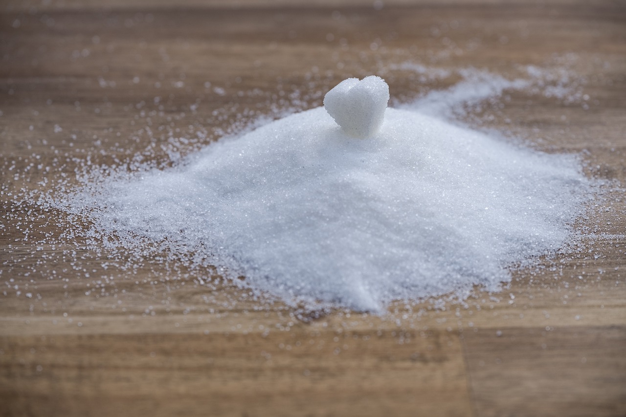 Small pile of granulated sugar on wooden surface with single heart-shaped sugar cube placed on top.
