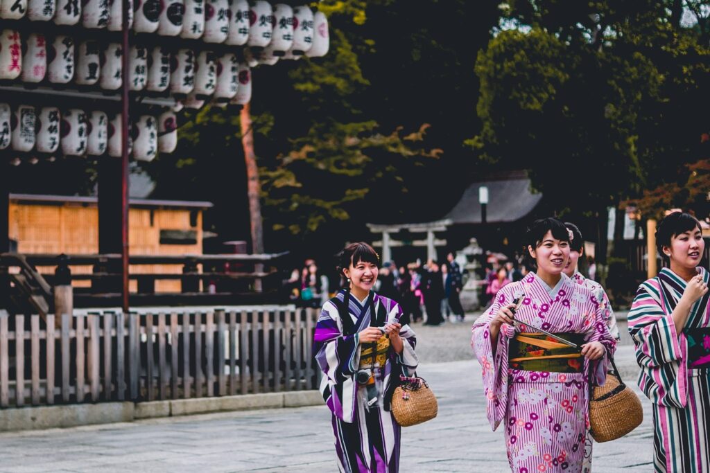Women in kimonos smiling as they walk past a traditional Japanese temple decorated with lanterns.