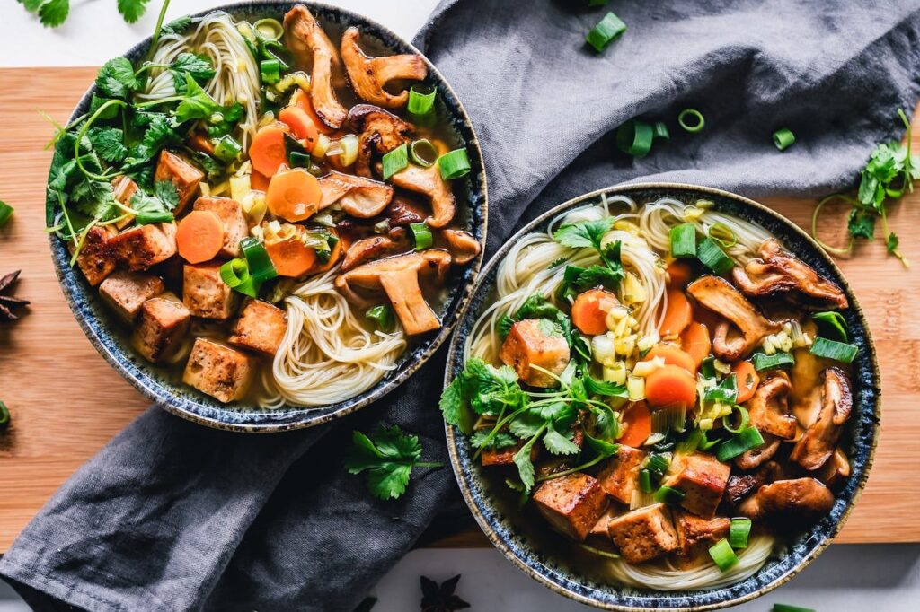 Two bowls of flavorful noodle soup garnished with mushrooms, tofu, carrots, and fresh greens.