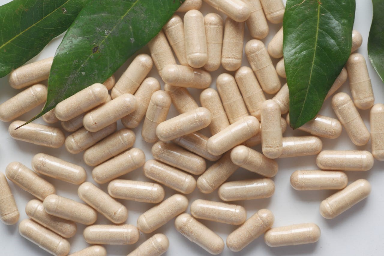 A close-up of beige supplement capsules scattered next to green leaves, symbolizing natural or plant-based supplements.