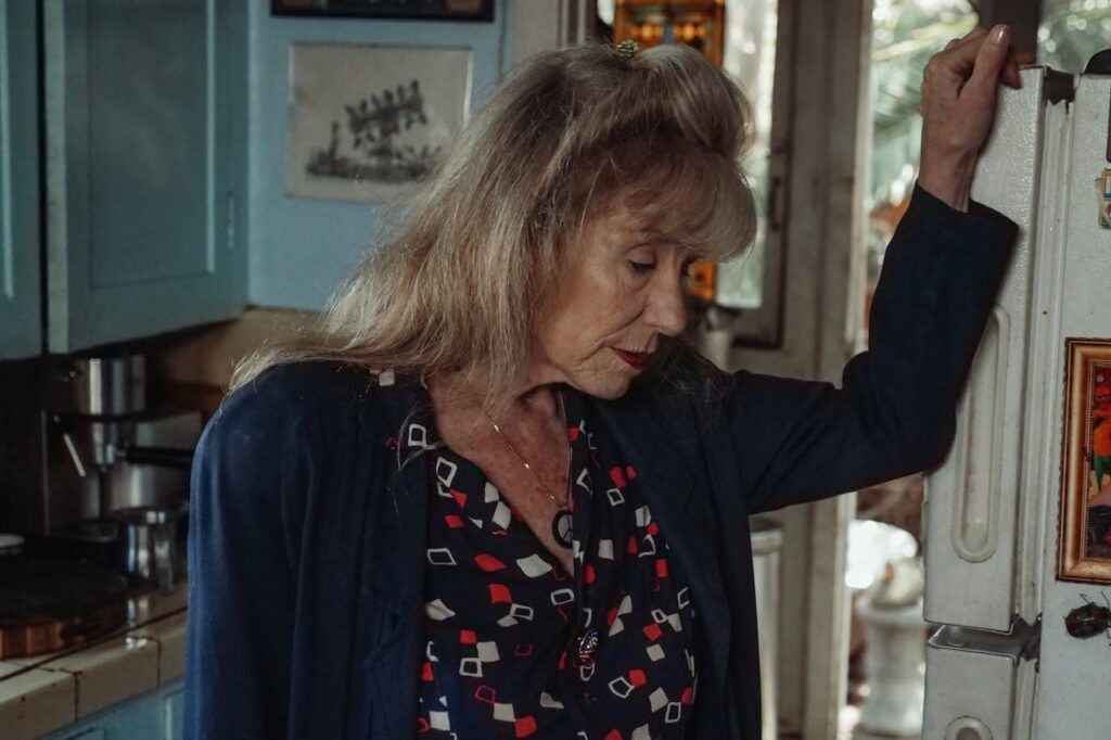 Older woman in a kitchen leaning against a refrigerator.