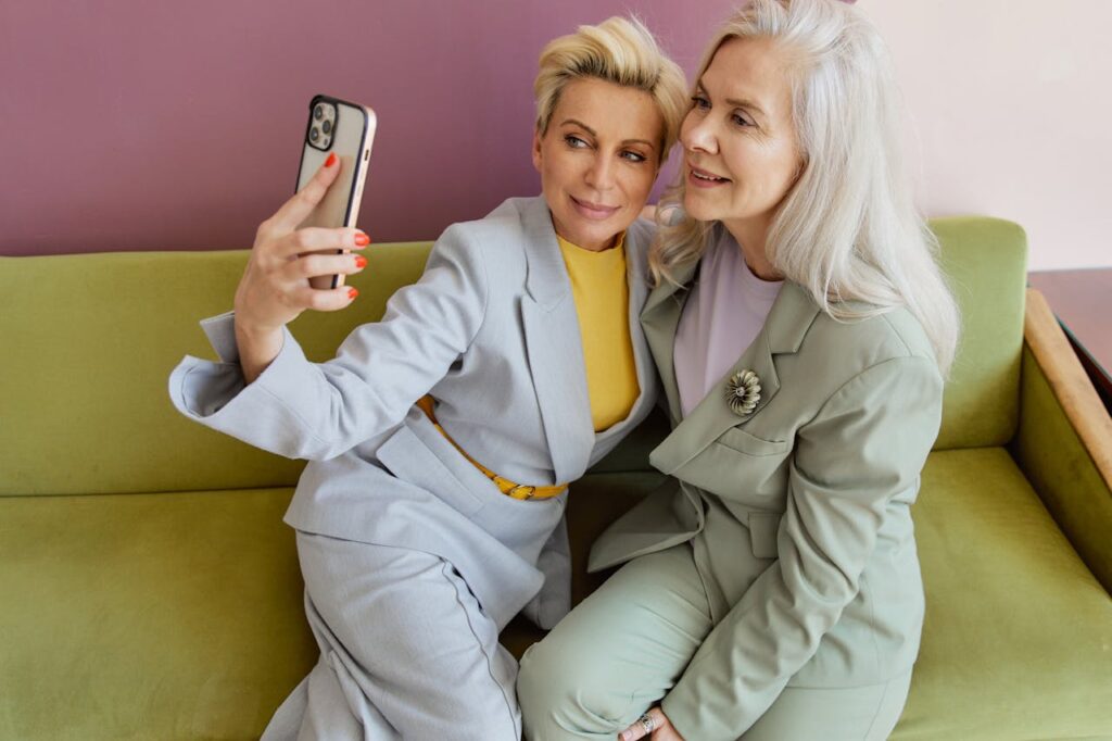 Two middle-aged women in suits sitting on a green sofa, smiling and taking a selfie together.
