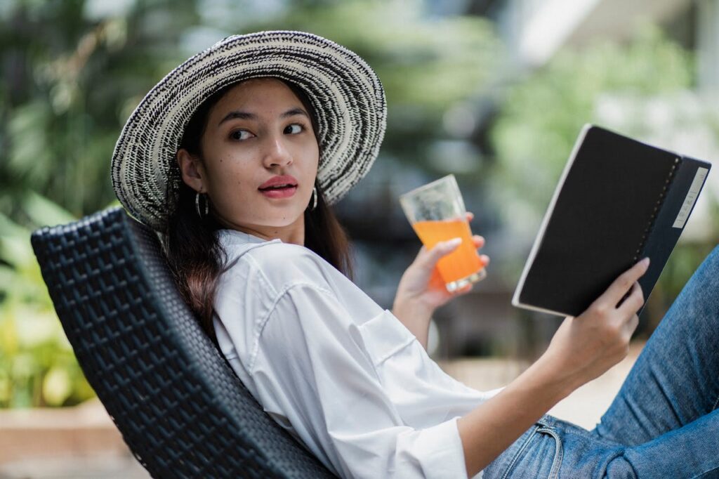 Woman relaxing outdoors with a notebook and drink, wearing a wide-brimmed hat.