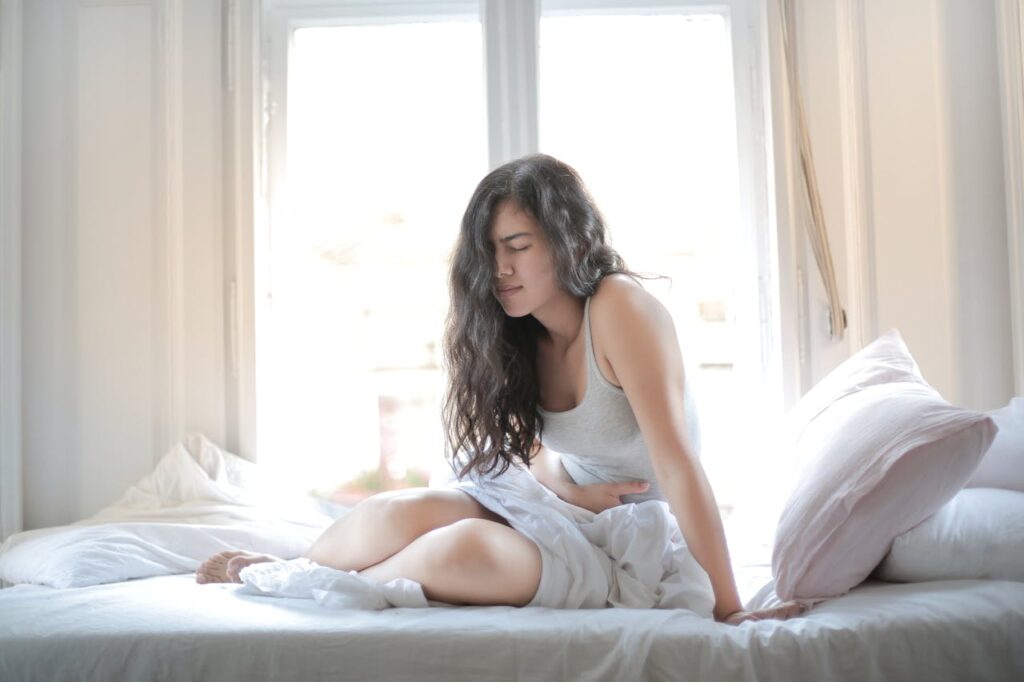 Woman sitting on a bed, holding her stomach, experiencing menstrual pain.