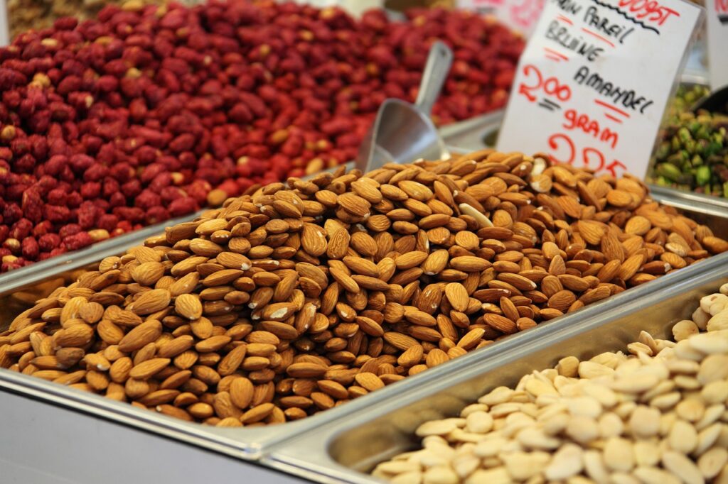 A pile of fresh almonds displayed in a market, surrounded by other types of nuts and dried fruits.