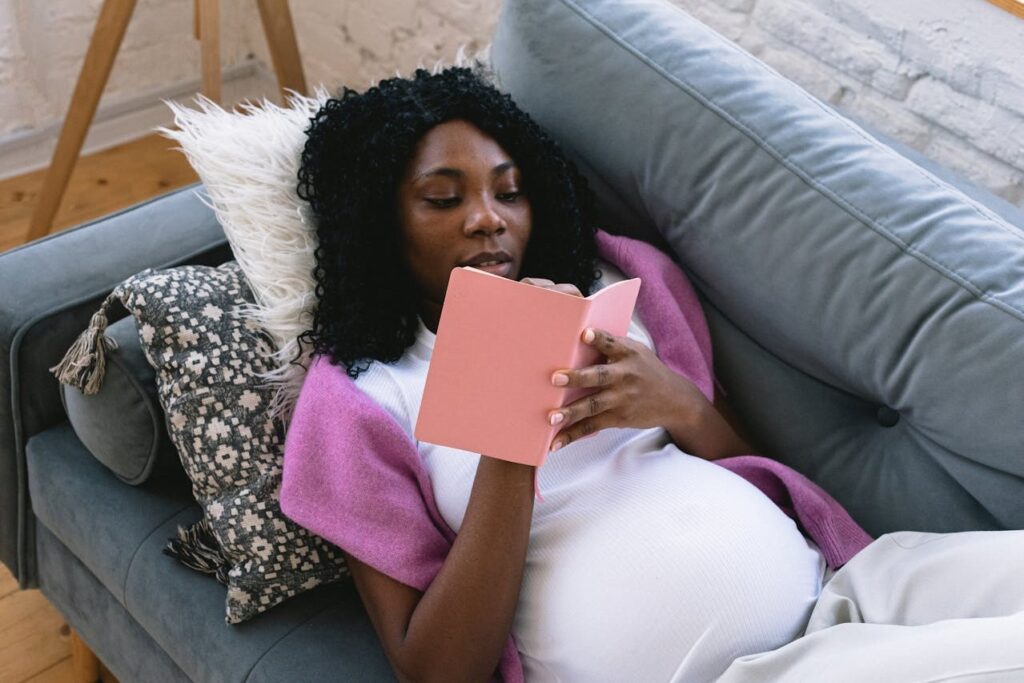 A pregnant woman in a lilac cardigan on a couch, reading a pink book.