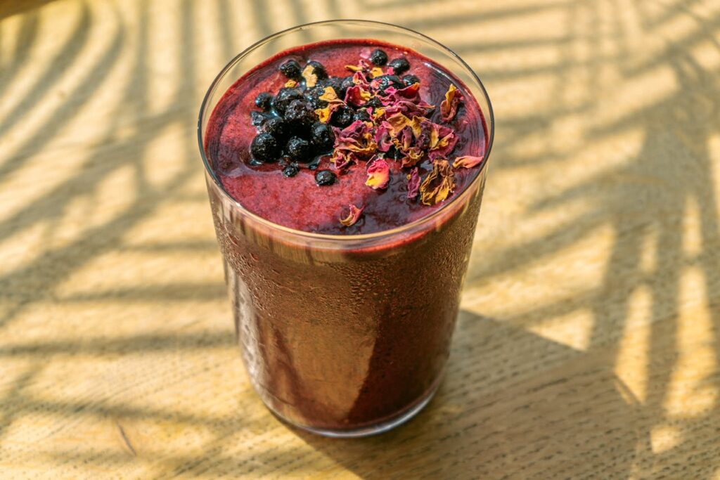 A purple smoothie topped with berries and flower petals, served in a clear glass.