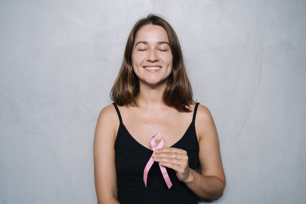 Smiling woman holding a pink breast cancer awareness ribbon with her eyes closed.